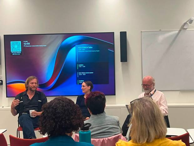 Matt speaking on a panel discussion at the University of Oxford, on the left hand side in black shirt and jeans, next to a woman and a man, in front of an audience. He speaks animatedly.